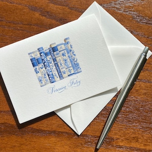 May include: White stationery set featuring a blue and white illustration of a stack of books. The design includes floral and patterned details. The name "Veronica Foley" is written in cursive below the books. A silver pen rests on a wooden surface.