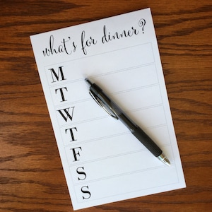 May include: A white sheet of paper with the text "what's for dinner?" written in black cursive. The paper has a list of days of the week, Monday through Sunday, written in black capital letters. A black pen is lying on the paper.
