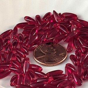 May include: A pile of red glass beads, some with a hole at one end, next to a US penny dated 2001.