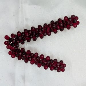 Può includere: Un fermaglio per capelli decorativo con un gruppo di piccole perline rotonde, rosso scuro. Le perline sono disposte in un motivo curvo a forma di vite, attaccate a una base di metallo argentato. Il fermaglio è progettato per tenere i capelli.