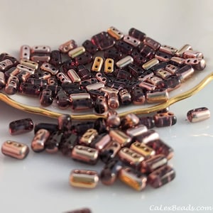May include: A pile of small, dark purple and gold glass beads. The beads are rectangular in shape and have two holes in each bead. The beads are scattered on a white surface.