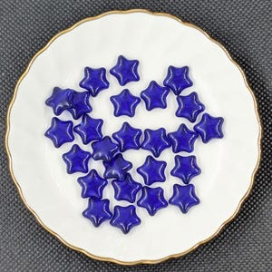 May include: A collection of deep blue star-shaped glass beads arranged on a white plate with a gold rim. The beads are evenly spaced, creating a visually appealing pattern. The plate is round and has a simple design.