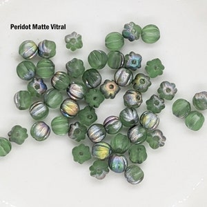 May include: A collection of green glass beads with a matte finish and iridescent accents. The beads are in various shapes, including round, oval, and flower-shaped. The beads are scattered on a white surface. Peridot Matte Vitral.