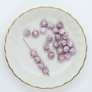 May include: A collection of pink and white striped glass beads on a white plate with a gold rim. The beads are arranged in a loose pile, with some beads strung on a thin wire.