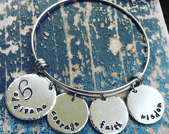 May include: Silver-toned bangle bracelet with four circular charms. The charms are stamped with the words "hope", "give me", "courage", "faith", and "wisdom". The bracelet is displayed on a distressed blue wooden surface.