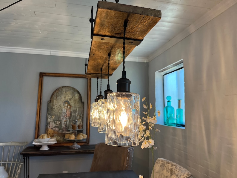 May include: A rustic wooden light fixture with four clear glass pendant lights hanging from black cords. The fixture is mounted to the ceiling and has a distressed wood finish. The lights are on and casting a warm glow.