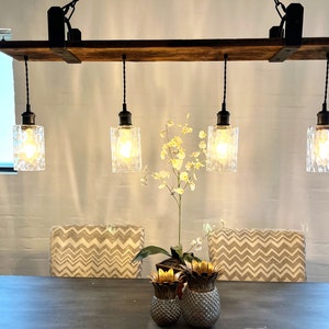 May include: A rustic wooden and metal light fixture with four clear glass pendant lights hanging from it. The fixture is mounted to the ceiling and has a black finish. The lights are turned on and illuminating the room.