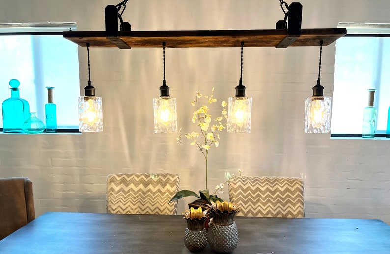 May include: A rustic wooden and metal light fixture with four clear glass pendant lights hanging from it. The fixture is mounted to the ceiling and has a black finish. The lights are turned on and illuminating the room.