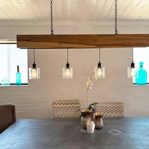 May include: A linear pendant light fixture featuring a long, rectangular natural wood beam suspended by two black chains. Four clear glass cylindrical shades hang from the beam, each containing an exposed filament bulb on a black cord.