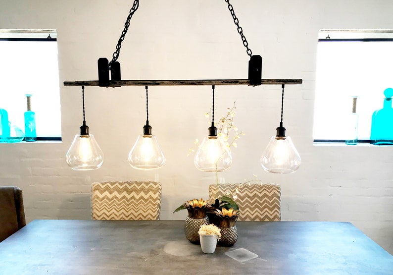 May include: A rustic wooden chandelier with four clear glass globe light fixtures hanging from black chains. The chandelier is suspended over a dining table with two chairs and a centerpiece of gold pineapple-shaped vases.