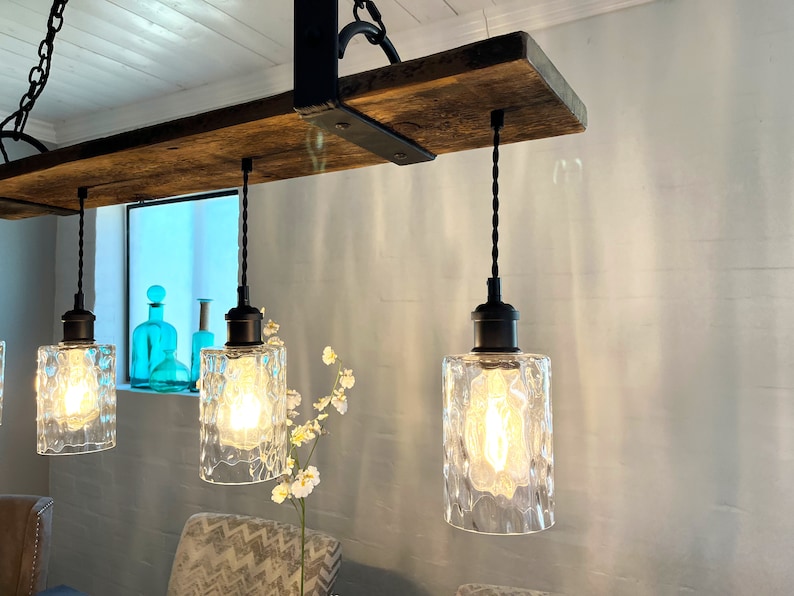 May include: A rustic wooden shelf with black metal brackets, supporting a chandelier with four clear glass pendant lights. The lights have a textured, frosted finish and are hanging from black cords.