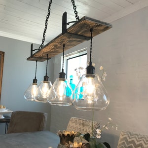May include: A rustic farmhouse style chandelier with five clear glass light bulbs hanging from a wooden beam. The beam is supported by black metal brackets. The chandelier is lit and casting a warm glow.