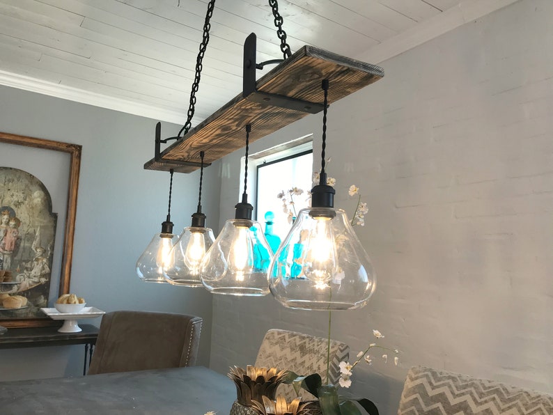 May include: A rustic farmhouse style chandelier with five clear glass light bulbs hanging from a wooden beam. The beam is supported by black metal brackets. The chandelier is lit and casting a warm glow.