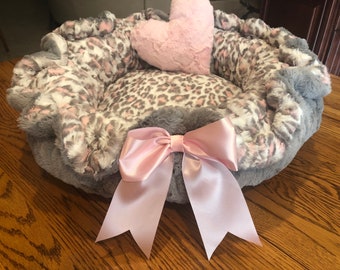 May include: A plush pet bed with a grey and leopard print design. The bed features a pink satin bow and a pink heart-shaped pillow. The bed is designed for small pets.