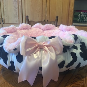 May include: A pink and white cow print pet bed with a large pink satin bow. The bed is made of soft, plush fabric and is perfect for small pets.