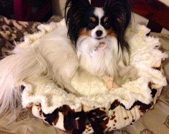 May include: A white and black papillon dog is lying in a cream-colored dog bed with a brown and white patterned base. The bed has a ruffled edge and a brown bow.
