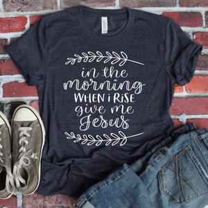 Puede incluir: Una camiseta azul oscuro con texto blanco que dice "in the morning when I rise give me Jesus" con un dise&ntilde;o floral arriba y abajo del texto.