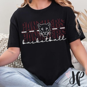 Puede incluir: Camiseta negra con la palabra "Panthers" en rojo y blanco, repetida en la parte superior. Un gráfico de cabeza de pantera está centrado encima de la palabra "basketball" en fuente cursiva. La camiseta es de ajuste cómodo.