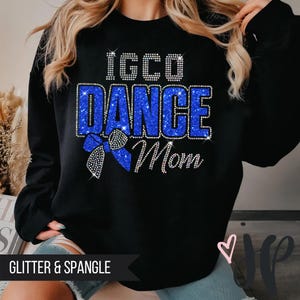 May include: Black sweatshirt with the text "IGCO DANCE Mom" in sparkling blue and silver rhinestones. A blue bow with rhinestone accents is below the words. The words "Glitter & Spangle" are at the bottom.