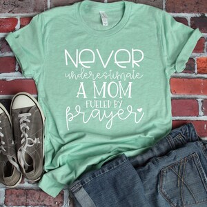 Peut inclure: Un t-shirt vert clair avec le texte "Never underestimate a mom fueled by prayer" en lettres blanches.