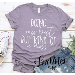 May include: A purple t-shirt with the text "Doing my best but kind of a mess" in white lettering. The shirt is on a white wooden background with a pair of white sneakers and a pair of blue jeans.