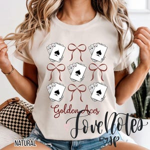 May include: A natural-colored t-shirt featuring a design of playing cards and bows. The design includes the text "Golden Aces" and "Love Notes" in a script font. The shirt is a casual style.