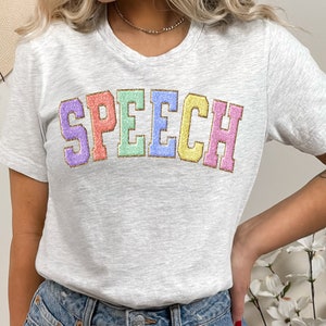 May include: A light gray t-shirt with the word "SPEECH" printed in rainbow colors with a gold outline.