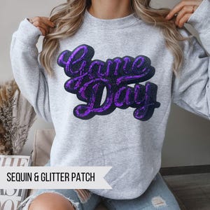 May include: Gray sweatshirt with a purple sequin and glitter patch that says "Game Day".