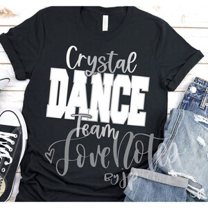 May include: Black t-shirt with white text that reads "Crystal Dance Team Love Notes by J." The shirt is on a white wooden surface with a pair of black sneakers and blue jeans.