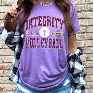 May include: Purple t-shirt with the text "Integrity Volleyball" in yellow and purple lettering. The shirt also features a white volleyball with a cross on it and the text "Play Purple Become Gold".