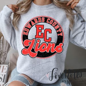 May include: A light gray sweatshirt featuring a distressed red and black graphic. The graphic includes the words "EDWARDS COUNTY" in a circle, "EC" in the center, and "Lions" in a script font. The sweatshirt has a crew neck.