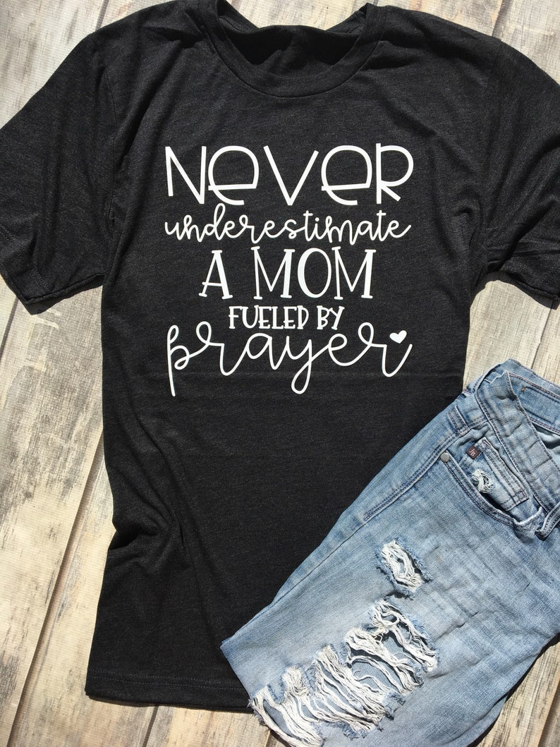 Peut inclure: Un t-shirt gris fonc&eacute; avec un texte blanc qui dit "Ne sous-estimez jamais une maman aliment&eacute;e par la pri&egrave;re" avec un petit symbole de c&oelig;ur.