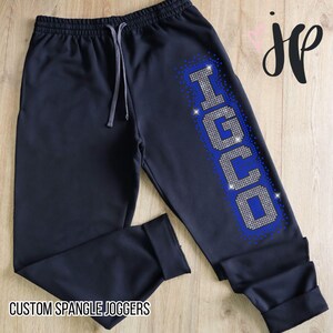 Pode incluir: Calças de jogging azul marinho com cintura com cordão e punhos nas tornozelos. A palavra "IGUO" é escrita com strass prateados contornados com strass azuis na perna esquerda. O texto "CUSTOM SPANGLE JOGGERS" está na parte inferior.