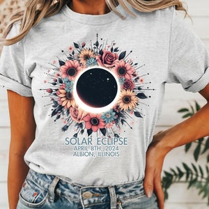 May include: A gray t-shirt with a floral design featuring a solar eclipse. The text on the shirt reads "Solar Eclipse April 8th, 2024 Albion, Illinois".