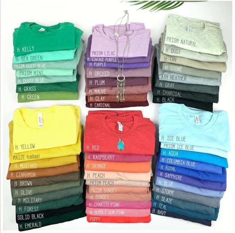 May include: A variety of t-shirt colors stacked in piles, with each pile labeled with a color name. The colors include Kelly, Sea Green, Dusty Blue, Mint, Grass, Green, Lilac, Vintage Purple, Purple, Orchid, Plum, Mauve, Clay, Cardinal, Natural, Dust, Tan, Stone, Heather, Gray, Charcoal, Black, Yellow, Maize, Mustard, Cinnamon, Brown, Olive, Military, Forest, Solid Black, Emerald, Red, Raspberry, Orange, Peach, Prism Peach, Sunset, Charity Pink, Bubble Gum Pink, Poppy, Ice Blue, Prism Ice Blue, Aqua, Columbia Blue, Royal, Sapphire, Blue, Storm, Slate, Teal, Navy.