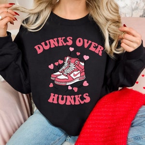 May include: Black sweatshirt with the text "Dunks Over Hunks" in pink letters. A pair of pink sneakers with white laces and a white swoosh logo are in the center of the text. Pink hearts are scattered around the sneakers.