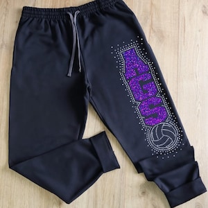 May include: Black sweatpants with a drawstring waist and cuffed ankles. The right leg features the word "SOUTH" in purple glitter, outlined with silver rhinestones, along with a rhinestone volleyball.