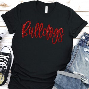 May include: A black short-sleeved t-shirt featuring the word Bulldogs written in a red glittery script font across the chest. The shirt is displayed alongside a pair of black and white low-top canvas sneakers and blue denim jeans with distressed details.