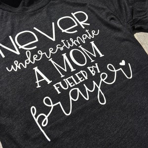 Peut inclure: Un t-shirt gris fonc&eacute; avec un texte blanc qui dit "Ne sous-estimez jamais une maman aliment&eacute;e par la pri&egrave;re" avec un petit c&oelig;ur blanc.
