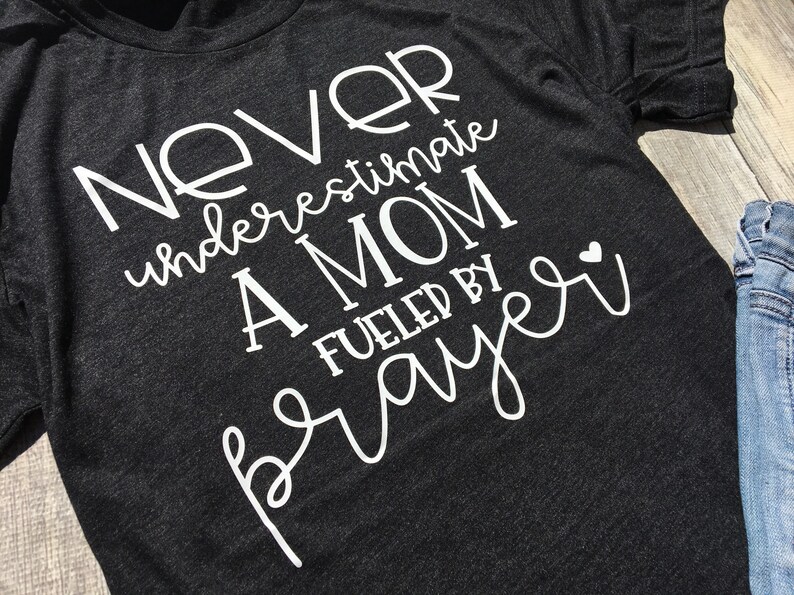 Peut inclure: Un t-shirt gris fonc&eacute; avec un texte blanc qui dit "Ne sous-estimez jamais une maman aliment&eacute;e par la pri&egrave;re" avec un petit c&oelig;ur blanc.