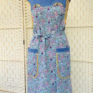 May include: A sleeveless apron featuring a light blue floral print with pink and purple flowers. It has a solid blue yoke with yellow trim, two front pockets with blue flaps and yellow piping, and a matching fabric tie at the waist.