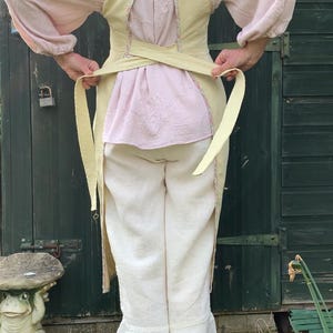 May include: A pale yellow apron with long ties is worn over a pink blouse and white trousers. The blouse has long, puffy sleeves and floral detailing. The trousers have a ruffled hem. The outfit is reminiscent of historical clothing.