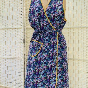 以下が含まれることがあります： 黄色いトリムとポケットが付いた花柄のラップエプロン。エプロンはマネキンに表示されています。生地は、暗い背景に青、紫、ピンクの花のパターンを特徴としています。エプロンはウエストで結ばれています。