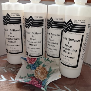 May include: Four white plastic squeeze bottles of Ridlen Adhesives Fabric Stiffener & Floral Porcelainizing Medium, each 16 fl. oz., positioned behind a small, square piece of fabric featuring a colorful floral pattern of roses and chrysanthemums.