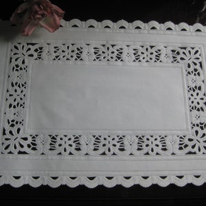 May include: A white rectangular placemat with a scalloped edge and intricate cut-out floral designs. The placemat has a plain center and a decorative border. A faded pink rose rests in the upper left corner.