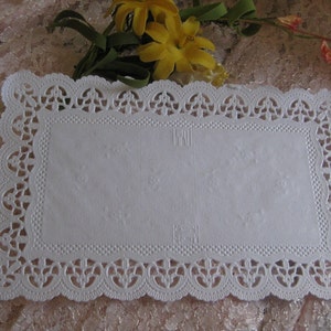 May include: White paper doily with a delicate lace pattern around the edges. The doily is rectangular and has a smooth, white surface.