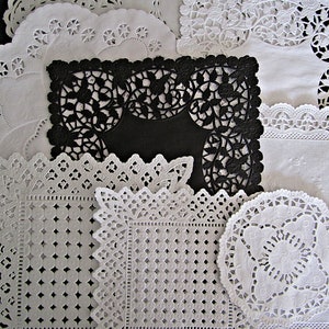 May include: A collection of white and black paper doilies in various shapes and sizes. The doilies feature intricate lace-like cut-out designs, including floral and geometric patterns. One black doily is rectangular, while others are round or square.