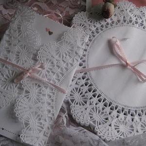May include: A white invitation with intricate lace detailing and a pink ribbon, alongside a matching doily. The invitation features a decorative lace border and is tied with a delicate pink ribbon. The overall theme is romantic and elegant.