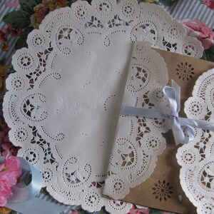 May include: Two white paper doilies with intricate lace patterns, one on top of a brown paper envelope tied with a light blue ribbon.