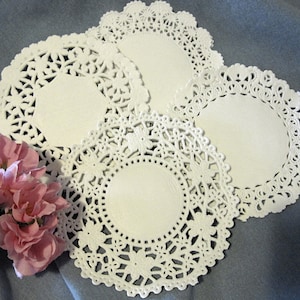 May include: Four white paper doilies with intricate lace designs. The doilies are arranged in a stack, with the largest doily on the bottom and the smallest doily on top.